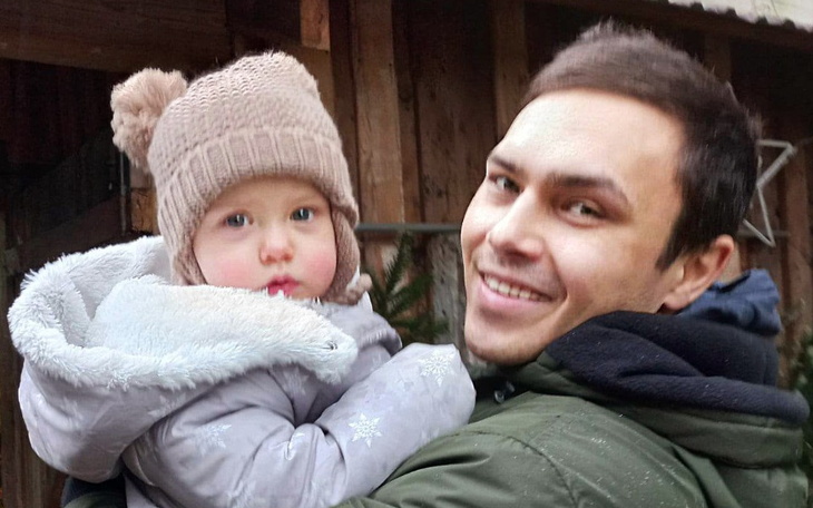 Mężczyzna trzymający małe dziecko w zimowej czapce na tle drewnianego budynku.