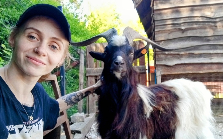 Osoba z kozą na tle drewnianej chaty, uśmiechnięta i zrelaksowana.