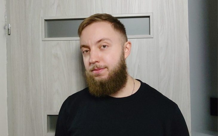 Mężczyzna z brodą stoi przed drzwiami, uśmiechając się do kamery.