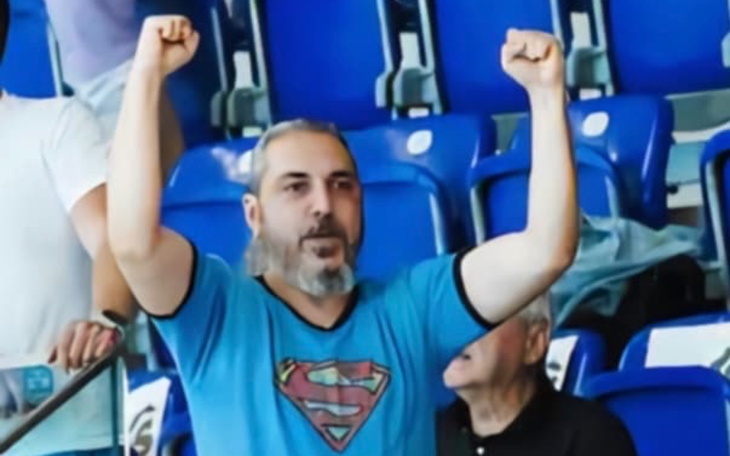 Kibic w niebieskiej koszulce z uniesionymi rękami na trybunach.