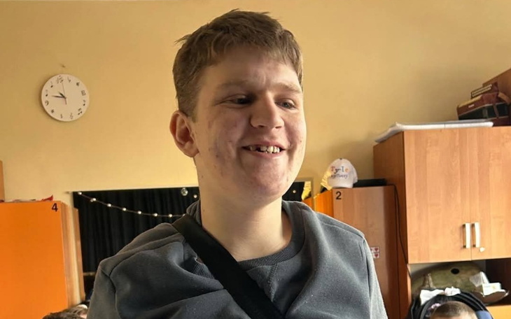 Młody chłopak uśmiechający się w klasie, otoczony rówieśnikami.