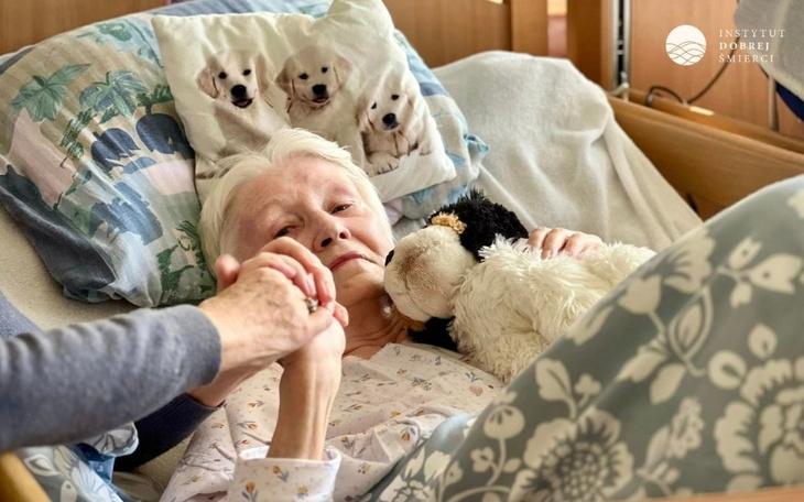 Starsza kobieta w łóżku, trzymająca rękę bliskiej osoby.