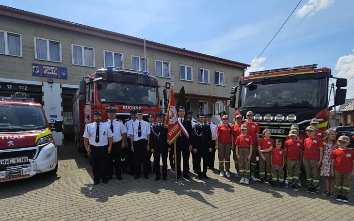 Grupa strażaków z dziećmi przed wozami strażackimi.