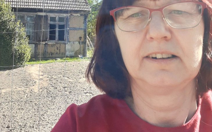 Kobieta w czerwonej koszulce, pozująca przed starym domem.