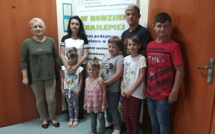 Grupa dzieci i dorosłych pozująca przed plakatem.