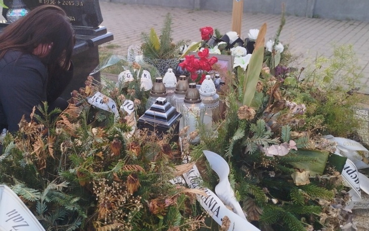 Osoba przy grobie, otoczona kwiatami i zniczami.