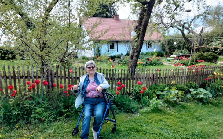 Starsza kobieta w ogrodzie, siedząca na wózku inwalidzkim, otoczona kwiatami.
