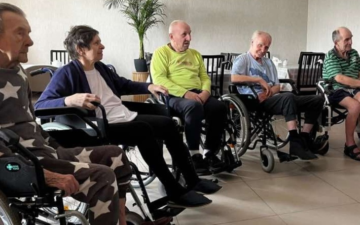 Grupa starszych osób siedzi na wózkach inwalidzkich w jasnym pomieszczeniu.