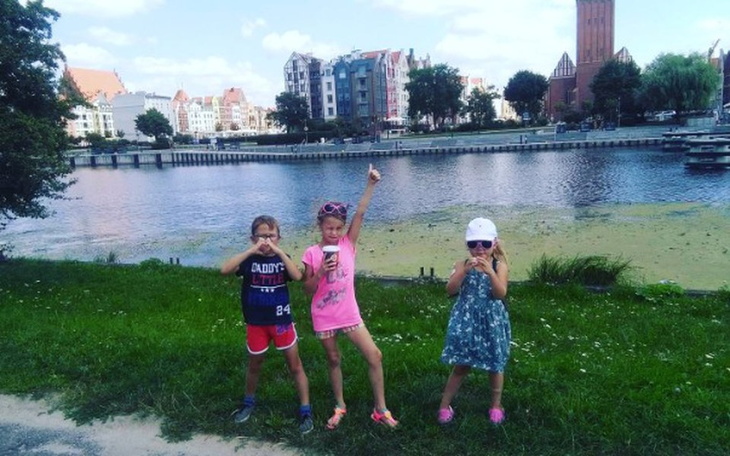 Trzy dziewczynki pozują na tle rzeki i miasta, ciesząc się słonecznym dniem.