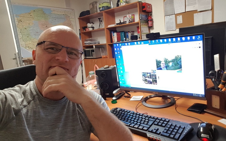 Mężczyzna w okularach przy biurku, z komputerem i mapą w tle.