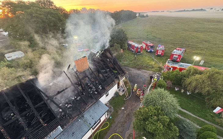 Zniszczony dach budynku po pożarze, w tle wozów strażackich.