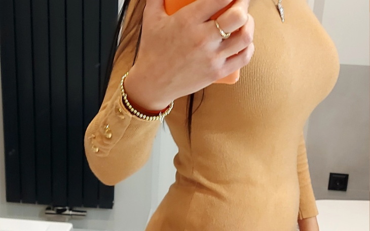 Kobieta w beżowej sukience, trzymająca telefon w lustrze.