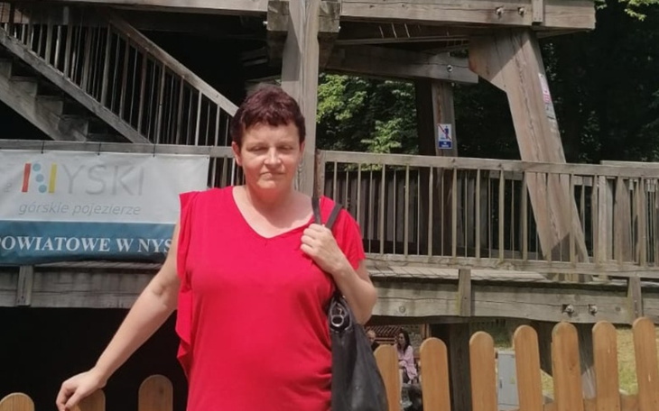 Kobieta w czerwonej bluzce na tle drewnianej konstrukcji.