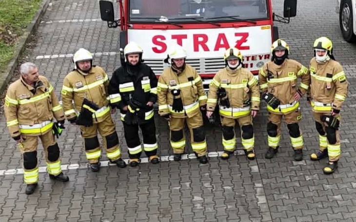Setka strażaków w żółtych mundurach przed czerwonym wozem straży pożarnej.