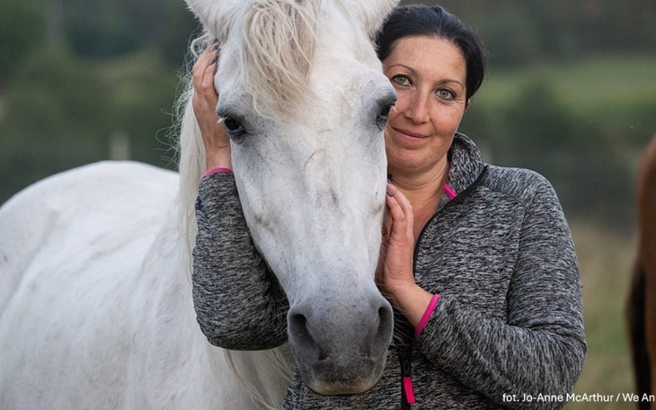 Kobieta przytulająca białego konia na tle zielonego krajobrazu.