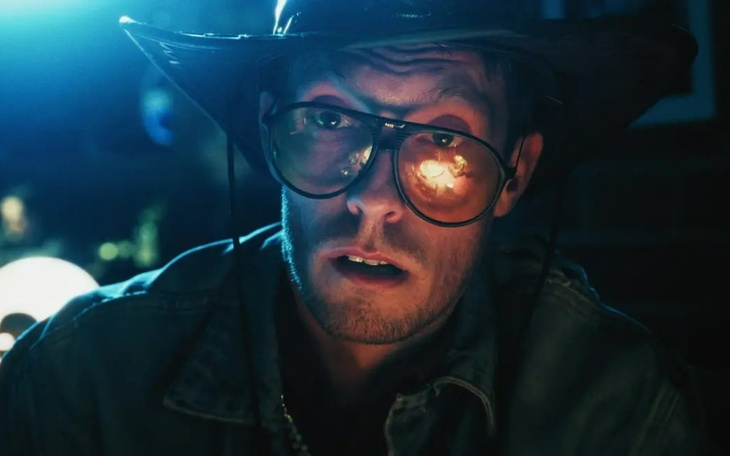 Mężczyzna w okularach i kapeluszu zaskoczony w ciemnym pomieszczeniu.