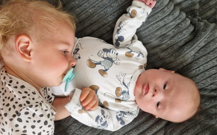 Dwoje małych dzieci leżących obok siebie na szarym kocu.