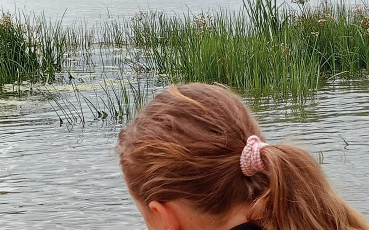 Kobieta z długimi włosami nad wodą, w tle roślinność.