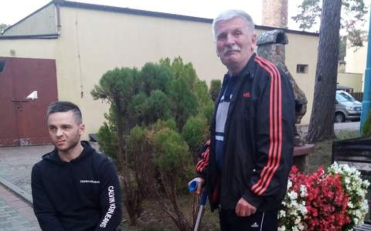 Mężczyzna w dresie z uśmiechem, siedzący obok drugiego mężczyzny w parku.