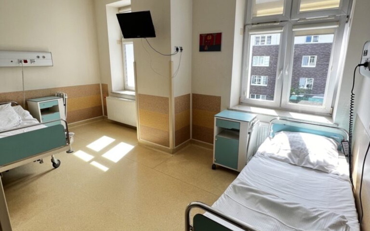 Pusty pokój szpitalny z dwoma łóżkami i telewizorem na ścianie.
