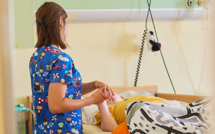 Pielęgniarka trzymająca rękę pacjenta w szpitalu, w tle łóżko szpitalne.