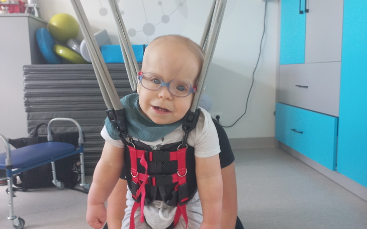 Małe dziecko w uprzęży terapeutycznej, uśmiechnięte i z okularami.