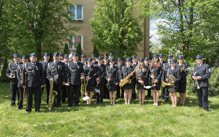 Orkiestra dęta w pełnym składzie, pozująca na tle zieleni.