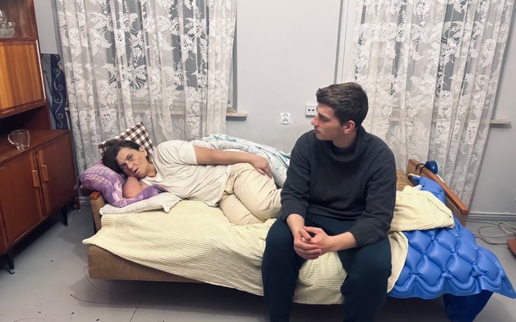 Osoba leżąca na łóżku, obok siedzi inna osoba, w tle zasłony.
