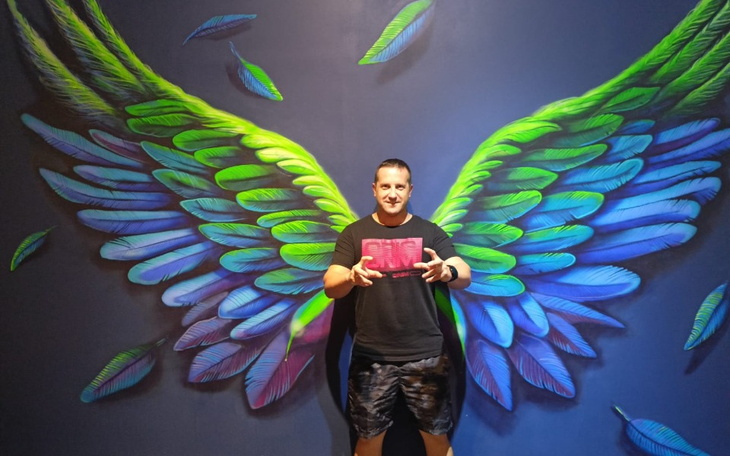 Mężczyzna pozujący przed muralem z kolorowymi skrzydłami.