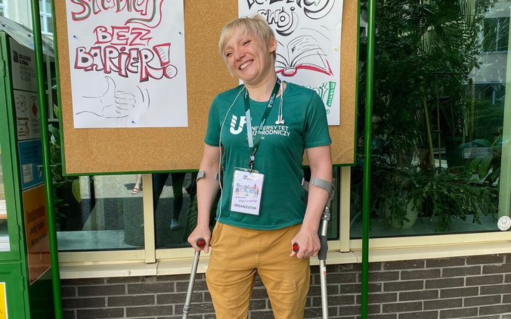 Osoba z uśmiechem, trzymająca kule, na tle tablicy z napisem 'Bez Barier'.