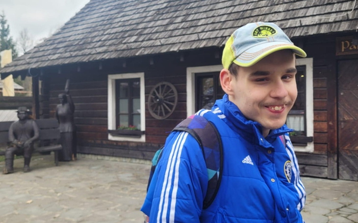 Młody mężczyzna w niebieskiej kurtce, uśmiechający się na tle chaty.