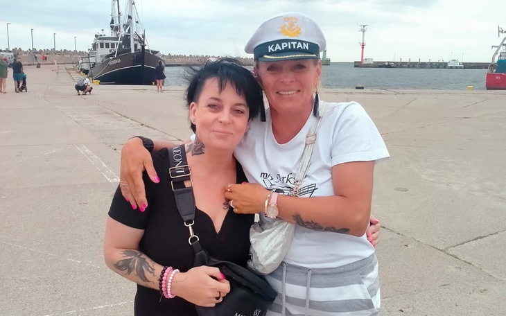 Dwie kobiety przytulają się na tle portu, jedna w kapitańskiej czapce.