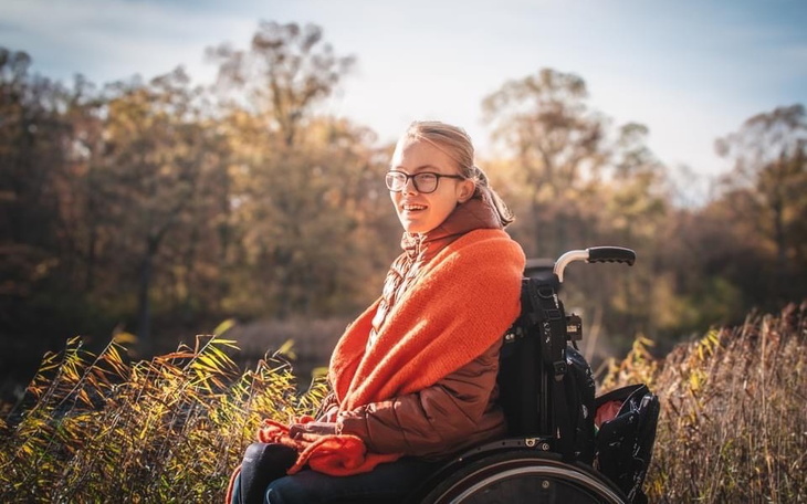 Młoda kobieta w wózku inwalidzkim, uśmiechająca się w otoczeniu natury.