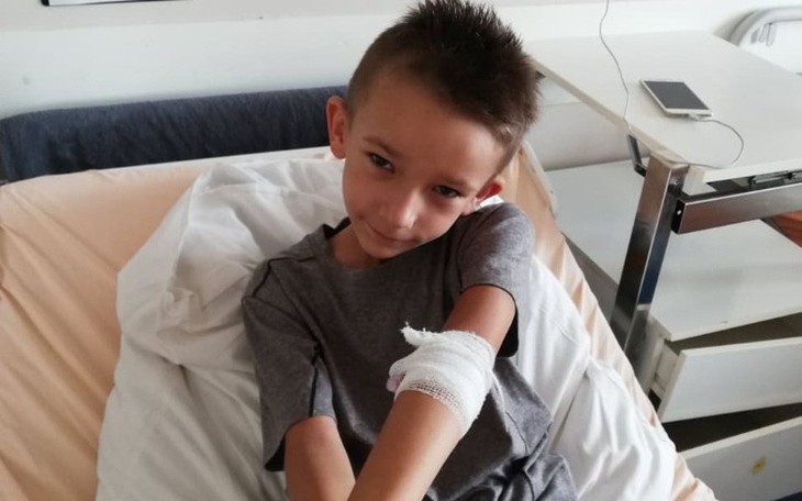 Chłopiec w szpitalu z opatrunkiem na ręce, uśmiechający się do kamery.