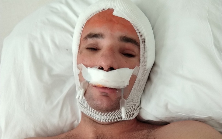 Mężczyzna w łóżku z opatrunkiem na głowie, wygląda na rekonwalescenta.