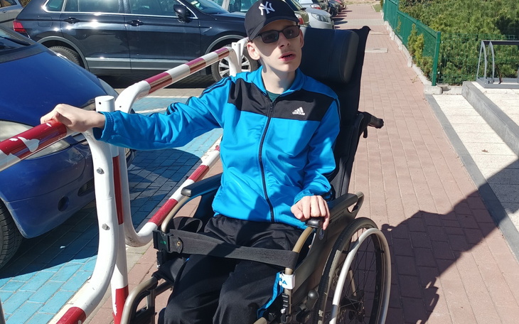 Chłopiec w wózku inwalidzkim, na zewnątrz, w niebieskiej kurtce.