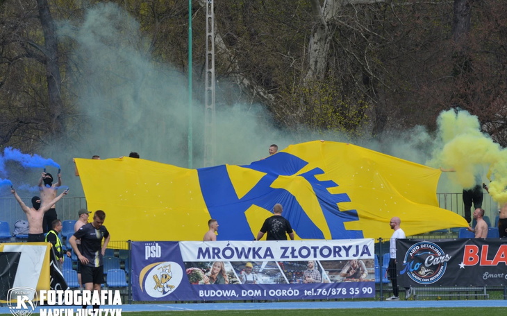Fani na stadionie z dużą flagą i kolorowymi dymami.