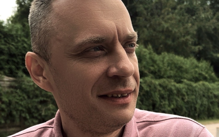 Mężczyzna w różowej koszuli, uśmiechający się w plenerze.