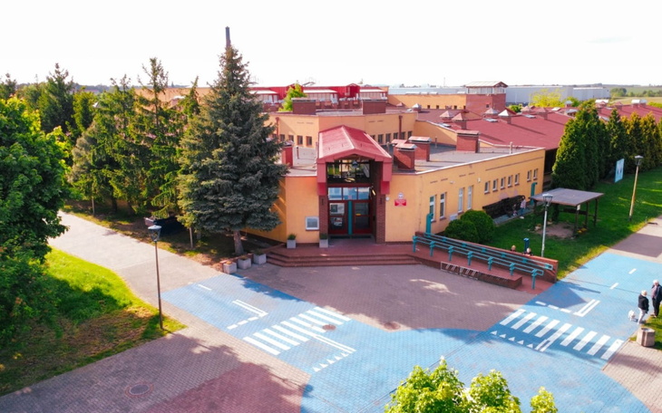 Widok na szkołę z zielonym otoczeniem i parkingiem.