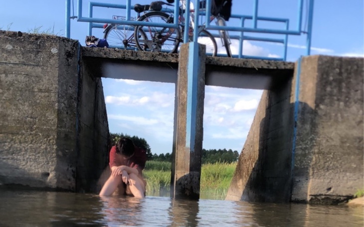 Osoba siedząca w wodzie, z rowerem w tle, otoczona zielenią.