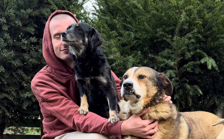 Mężczyzna z dwoma psami, uśmiechając się w otoczeniu zieleni.