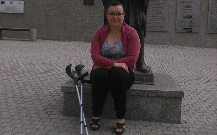 Kobieta siedząca na ławce z kulami, przy pomniku.