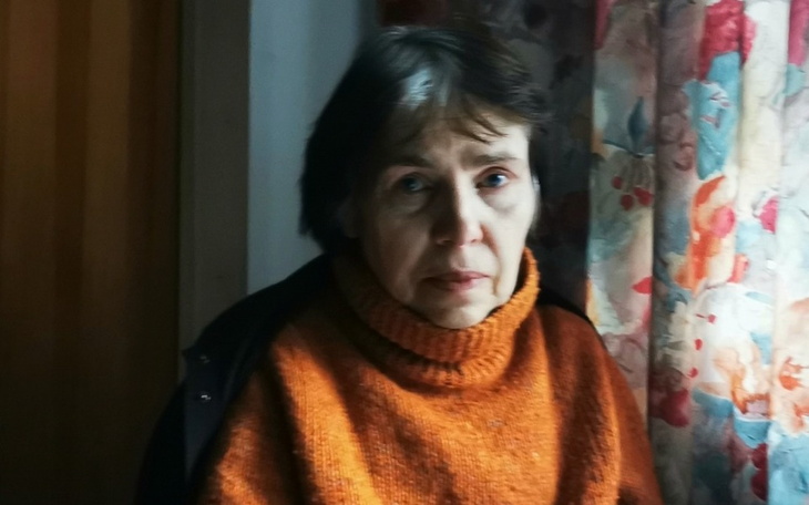 Portret starszej kobiety w pomarańczowym swetrze, z zamyślonym wyrazem twarzy.