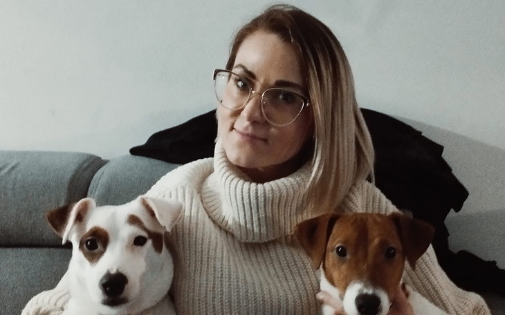 Kobieta z dwoma psami na kanapie, uśmiechnięta i w swetrze.