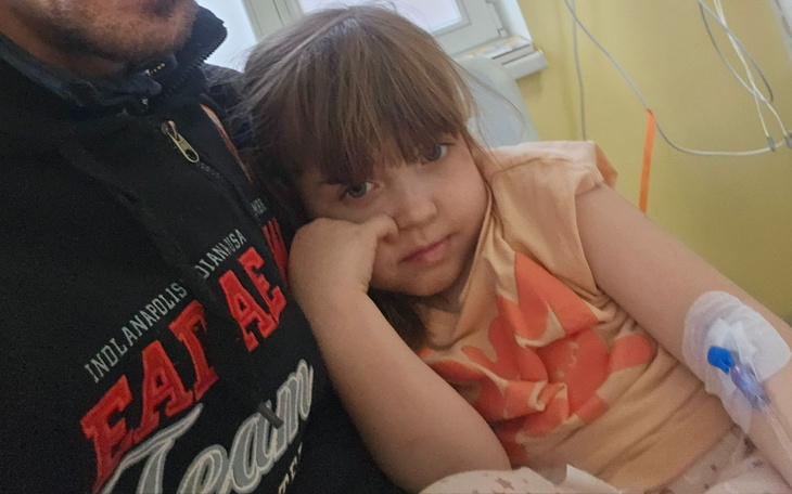 Dziewczynka w szpitalu, z opatrunkiem na ręce, wygląda na zamyśloną.