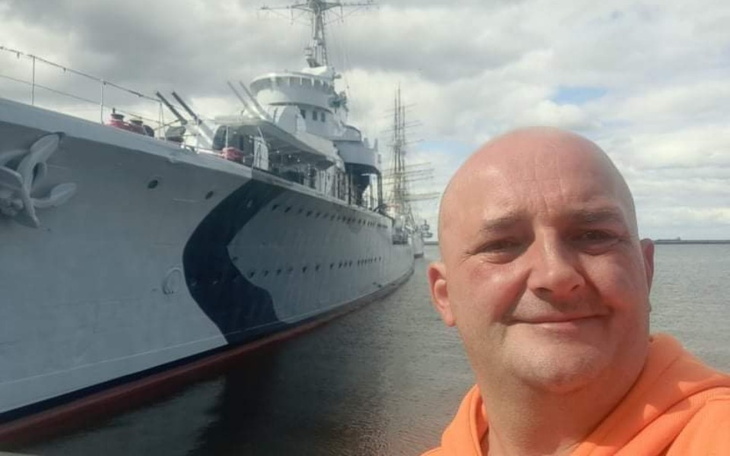 Mężczyzna pozujący z dużym statkiem w tle, nad wodą.