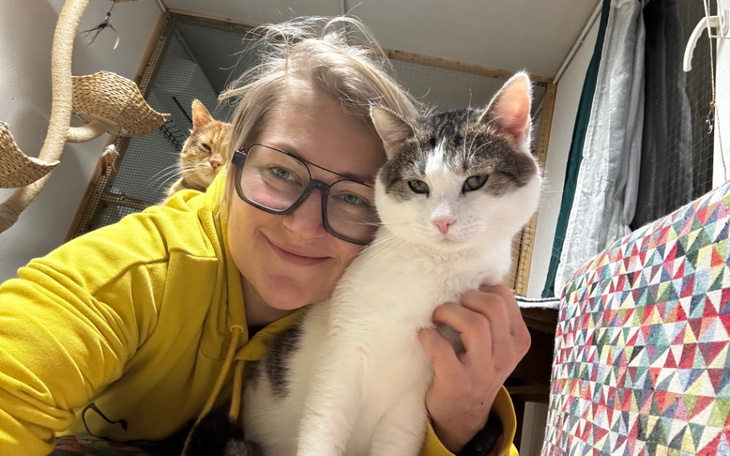 Kobieta z okularami przytula kota, w tle widać innego kota.