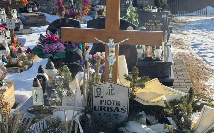 Grób z krzyżem i zniczami, otoczony kwiatami, w zimowej scenerii.