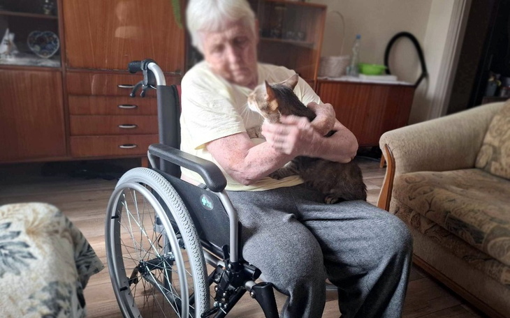 Starsza kobieta w wózku inwalidzkim przytula kota w domowym otoczeniu.