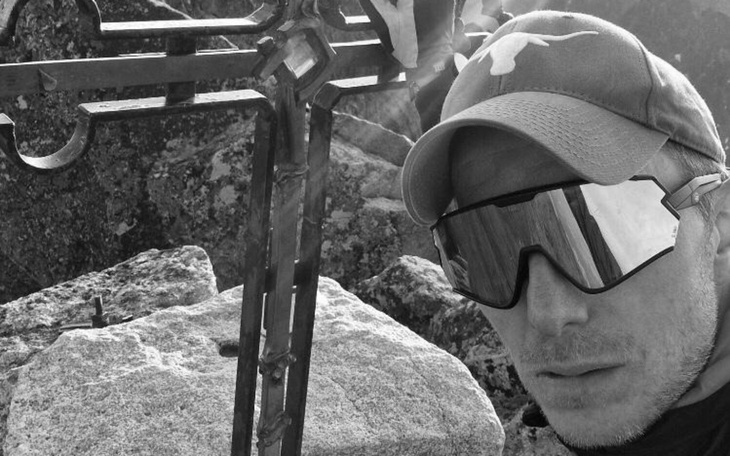 Mężczyzna w okularach przeciwsłonecznych przy metalowym krzyżu na szczycie góry.